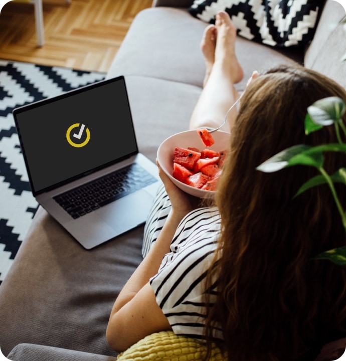 Kvinde ligger på en sofa og spiser en skål med frugt med en bærbar computer ved siden af sig med Norton-logo
